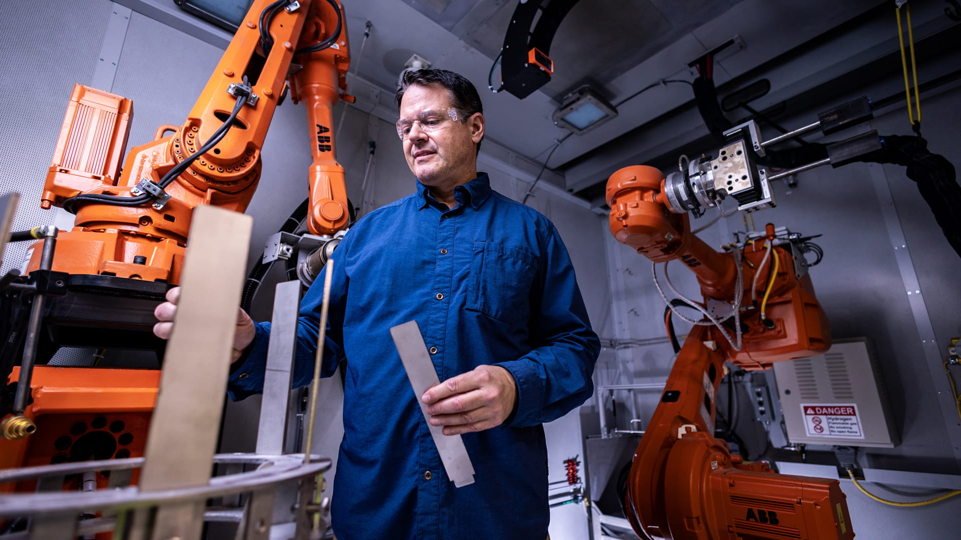 Middle-aged adult male wearing safety glasses and a button-down collared shirt operating orange machinery. 