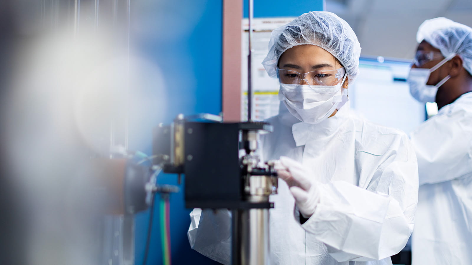 Woman wearing a white hair net, white mask, white rubber gloves, white lab coat and safety glasses working in a lab.
