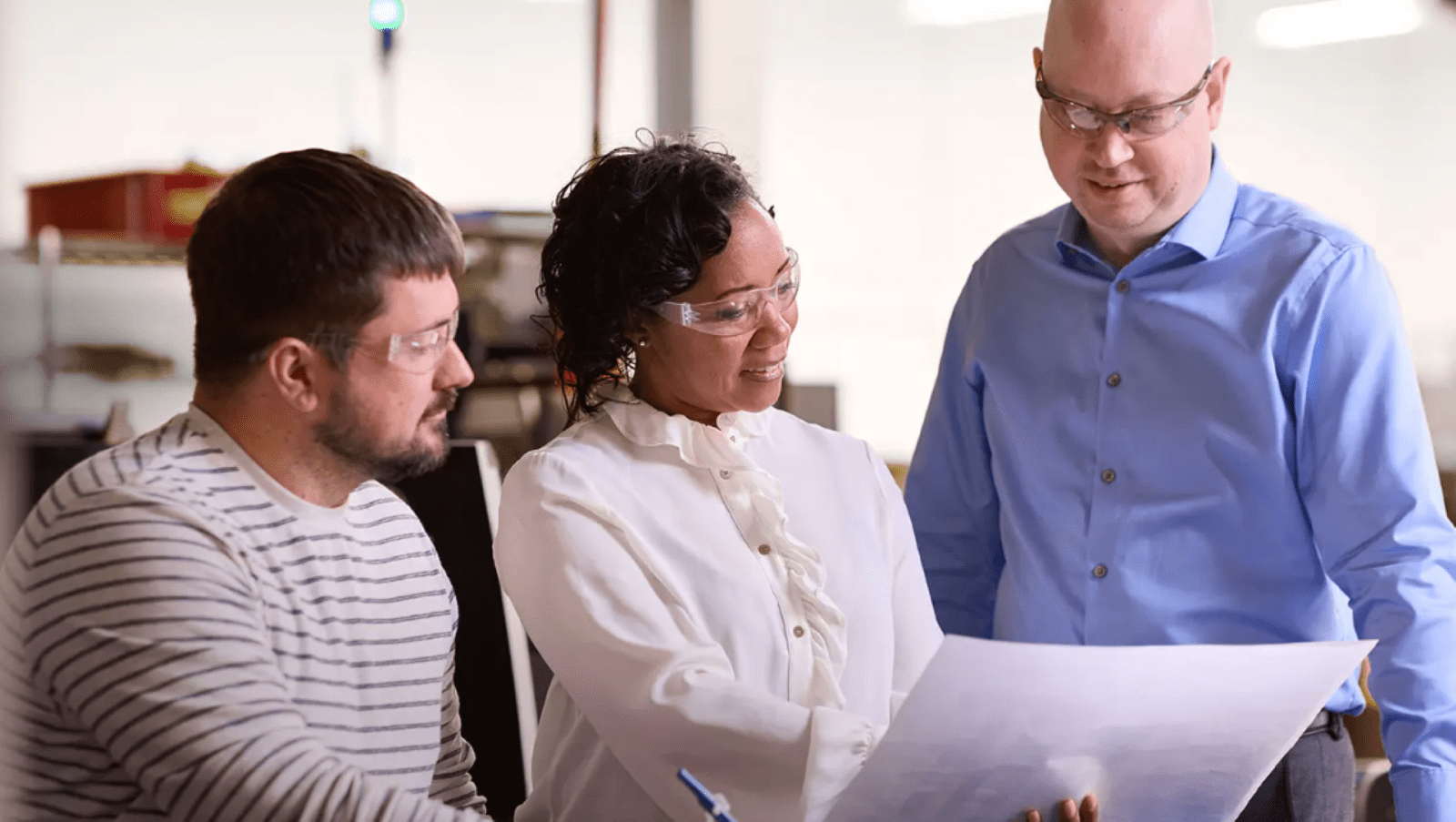 Adult female pointing out information on a large sheet to two adult males - all wearing safety glasses - in an office setting.